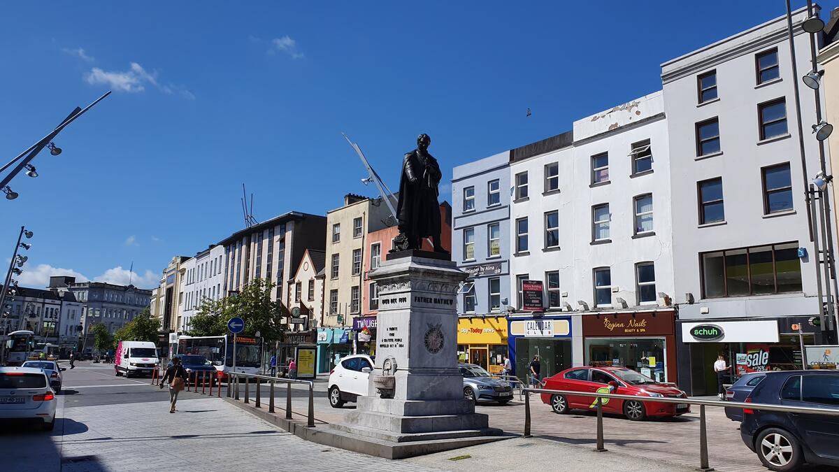 The Father Mathew statue on St Patrick&rsquo;s Street in Cork city, with shops, traffic and pedestrians under a bright blue sky.